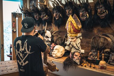 An artist works on makeup or costumes in a room filled with numerous demon and devil masks, heads, and props. | © Zell am See-Kaprun Tourismus