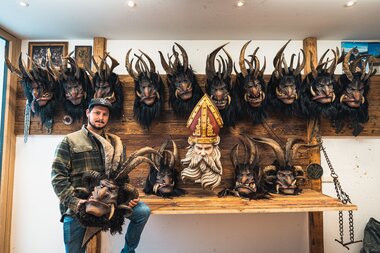 A person stands beside a wall displaying various Krampusköpfe with horns and a decorative red and gold hat. | © Zell am See-Kaprun Tourismus