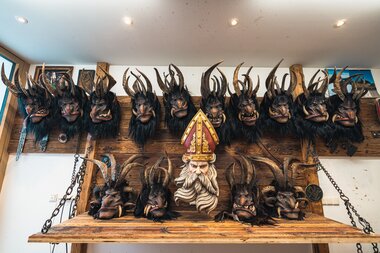 A collection of intricately crafted masks with horns, displayed on a wooden shelf, including a central figure resembling a bishop or saint. | © Zell am See-Kaprun Tourismus