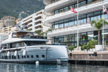 Yacht docked in harbor in front of a modern building with flags and palm trees. | © Studio F. A. Porsche