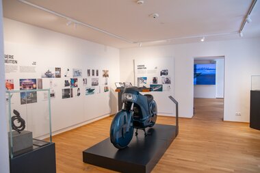 Exhibition with pictures and a sculpture of a motorcycle engine in a gallery space. | © Nikolaus Faistauer Photography