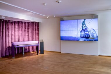 An empty exhibition room with a television displaying an aerial tramway and a piano in front of purple curtains. | © Nikolaus Faistauer Photography