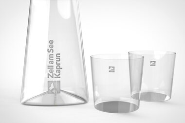 Glass carafe and two matching glasses with logos on a white background. | © Zell am See-Kaprun Tourismus