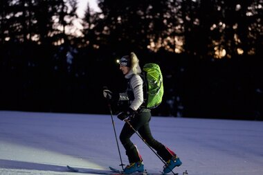 Skier entering a nighttime ascent on a remote snowy slope, surrounded by dark pine trees, with hiking gear and a headlamp. | © Zell am See-Kaprun Tourismus