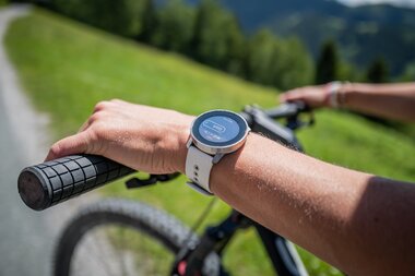Close-up of a wrist with a smartwatch on a mountain bike in a green landscape. | © Suunto Philipp Reiter