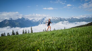 Person trail running in the Alps with mountain peaks and clouds in the background, surrounded by green meadow. | © Suunto Philipp Reiter