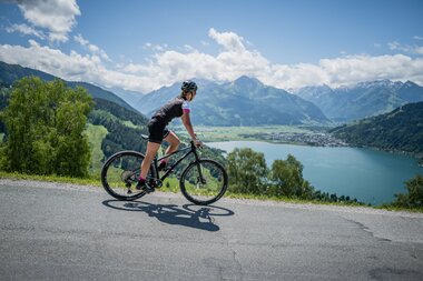 A person rides a mountain bike on a hilly road with a lake and mountains in the background. | © Suunto Philipp Reiter