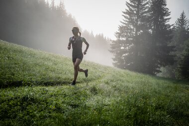 Person trail running on a grassy slope with trees in the background, morning mist, and sunlight. | © Suunto Philipp Reiter