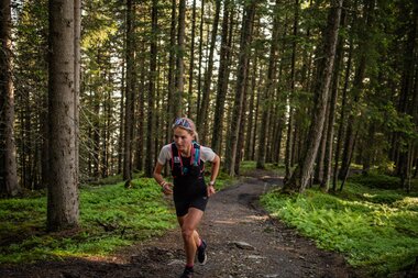 A person is running on a forest trail with trees and sunlight in the background, wearing running clothes and a backpack. | © Suunto Philipp Reiter