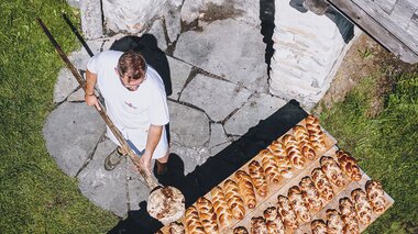 Baker baking loaves in an outdoor stone oven, surrounded by green grass. | © EXPA JFK