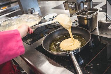 A Kaiserschmarrn is being cooked in a pan on the stove, with batter pouring into the pan, while someone assists in the kitchen. | © Zell am See-Kaprun Tourismus