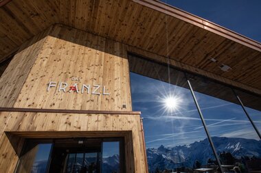 Modern wooden structure with large glass windows and a view of snow-covered mountains on a sunny day. | © Schmittenhöhe