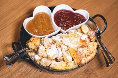 Kaiserschmarrn on a wooden tray, served with caramelized onions and two sauces in heart-shaped dishes, with a powdered sugar topping. | © Zell am See-Kaprun Tourismus
