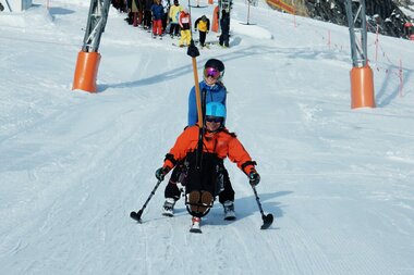 Skier user on an adaptive ski device on a snowy slope, surrounded by other skiers and a ski lift line. | © Up adaptive sports 