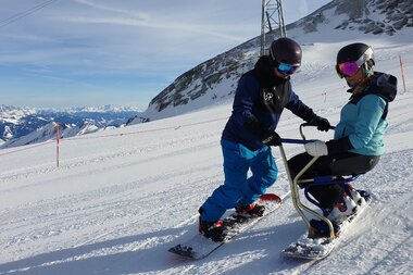 A person with a disability is learning to ski with assistance on a special ski device. Snow-covered mountains and a clear blue sky are in the background. | © Up adaptive sports 
