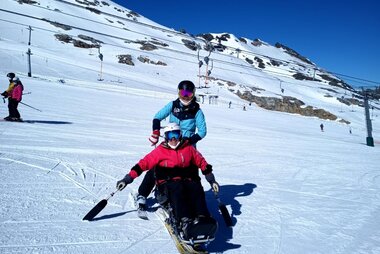 Person on skis on a snowy slope, accompanied by a supervisor in ski gear, under a clear blue sky. | © Up adaptive sports 