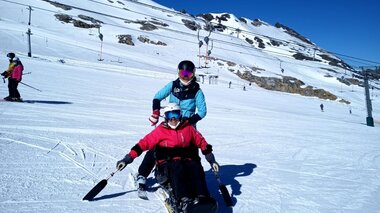 Person on skis on a snowy slope, accompanied by a supervisor in ski gear, under a clear blue sky. | © Up adaptive sports 