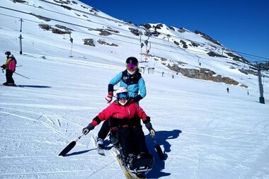 Person on skis on a snowy slope, accompanied by a supervisor in ski gear, under a clear blue sky. | © Up adaptive sports 