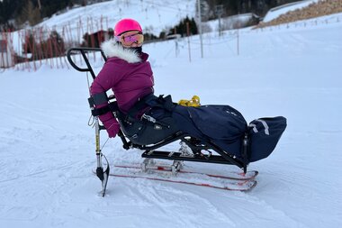 Person using adaptive skiing equipment on snow, wearing a pink helmet and goggles, dressed in winter clothing. | © Up adaptive sports 
