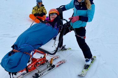 An individual participating in adaptive skiing is assisted by a companion on skis on a snowy slope. | © Up adaptive sports 