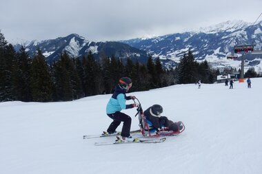 Two skiers on a snow slope, one pulling a wheelchair with an adaptive ski vehicle, with mountains and ski lift in the background. | © Up adaptive sports 