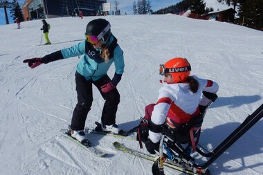 Two skiers on a slope, one using a sit-ski and receiving guidance from the other. | © Up adaptive sports 