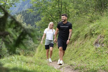 Two people hike on a narrow trail through a green mountain landscape with fir trees and mountains in the background. | © Zell am See-Kaprun Tourismus
