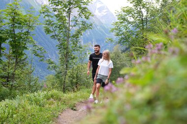Two people hike on a forest trail in a mountainous, green landscape with snow-capped peaks in the background. | © Zell am See-Kaprun Tourismus
