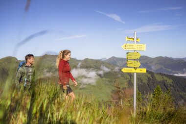 Two hikers walking on a mountain trail with mountain peaks in the background, signs with trail markers are visible. | © Schmittenhöhe