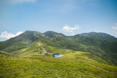 Landscape with green hills, mountains, and a small lake under a blue sky. | ©  Markus Landauer, Saalbach Hinterglemm