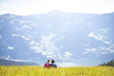 Two people sitting on a meadow with a view of Lake Zell and the surrounding mountains. | © Schmittenhöhe