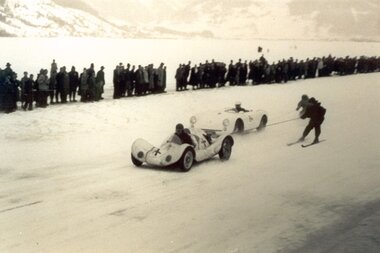 A racing car is being pulled on the snowy ice track as a person on skis guides it, with a crowd of spectators in the background. | © Zell am See-Kaprun