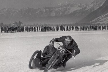 Historical ice speedway race with a motorcycle on icy terrain, background shows a large crowd and snow-covered mountains. | © ÖAMTC