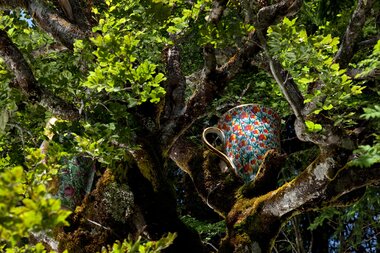 A colorful teapot hangs among the branches of a tree, surrounded by green leaves, as part of the Art on the Mountain exhibition. | © Schmittenhöhebahn