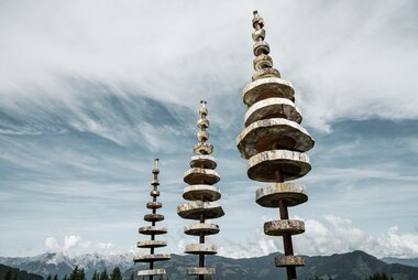 Art installation of multiple cylindrical sculptures on a mountain with a mountain range in the background and a cloudy sky. | © Schmittenhöhebahn