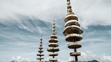Art installation of multiple cylindrical sculptures on a mountain with a mountain range in the background and a cloudy sky. | © Schmittenhöhebahn