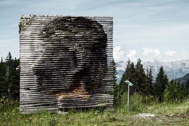 Wood sculpture of a portrait made visible through tomography technique, set against a mountain landscape with forest and mountains in the background. | © Schmittenhöhebahn