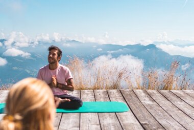 People practicing yoga on a wooden platform with mountain and sky background, one sitting cross-legged and the other standing on a yoga block. | © Johannes Radlwimmer
