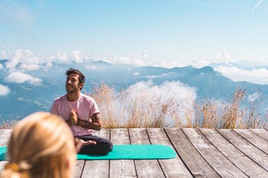 People practicing yoga on a wooden platform with mountain and sky background, one sitting cross-legged and the other standing on a yoga block. | © Johannes Radlwimmer