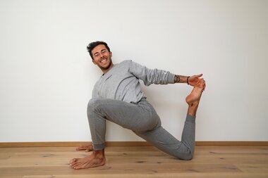 Person in a yoga pose in a bright room, smiling, sitting on the floor with one leg crossed and in a backward bend. Showing a radiant smile. | © Marcel Clementi