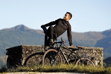 Person in a yoga pose on a large stone slab with a bicycle in front of mountain scenery outdoors. | © Marcel Clementi