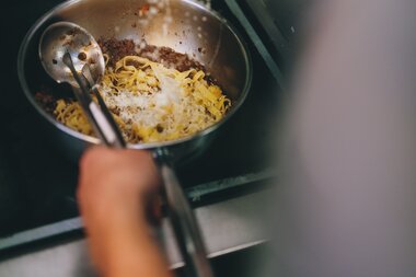 Person prepares pasta in a pan on a stove, adding salt. | © Zell am See-Kaprun Tourismus