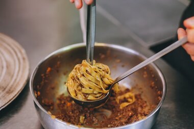 A hand holds scissors and a fork to transfer noodles from a pan in a professional kitchen. | © Zell am See-Kaprun Tourismus