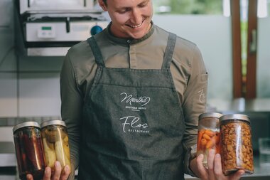 Man in kitchen holding three jars of pickled vegetables in each hand, wearing a apron with Flo's Restaurant logo. | © Zell am See-Kaprun Tourismus