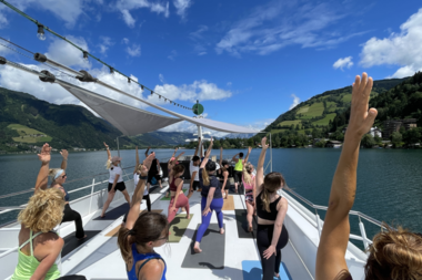 Yoga class on a boat with a view of a lake and surrounding mountains, clear blue sky.