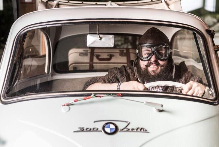 Person driving a vintage BMW 2002, wearing goggles and a hat, smiling. | © Fam. Voetter