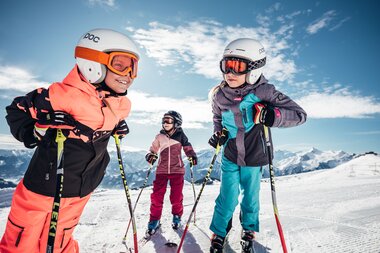 Children enjoying skiing lessons on Mount Schmitten, surrounded by snowy mountains and blue sky.