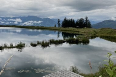 A peaceful lake view with surrounding mountains and dark clouds in the sky, with a wooden dock in the foreground. | © Stephanie Spatt I Zell am See-Kaprun Tourismus