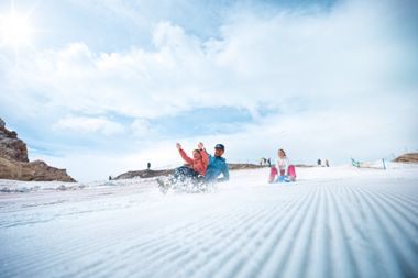 People sliding on a snowy slope outdoors, with mountain landscape in the background and a cloudy sky.