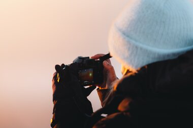 A person taking a photo with a camera during sunset, wearing a white hat and dark clothing. | © manueldietrichphotography